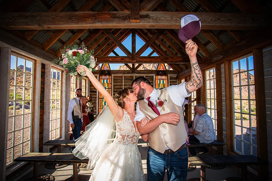 Wedding couple celebrating in a rustic chapel at Nelson Ghost Town, bride lifting her bouquet and groom raising his hat in warm golden light